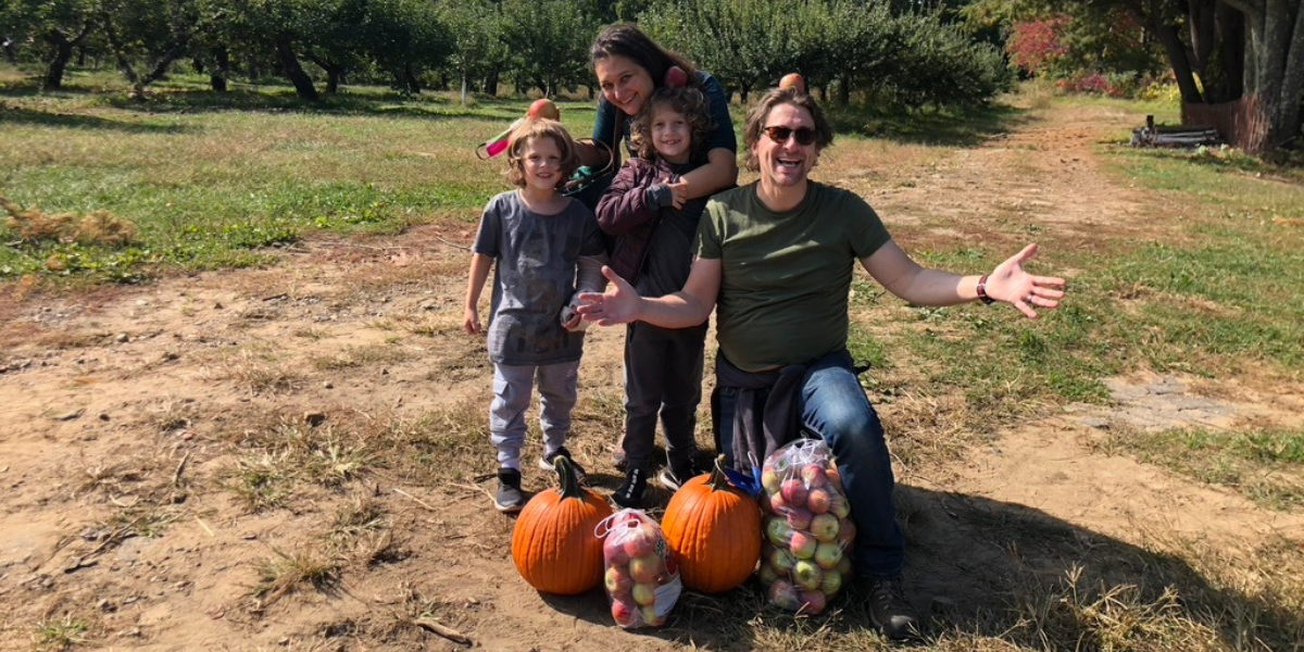 Ethan kneeling with an apple on his head, with his wife and 2 sons. Pumpkins and bags of apples in the front