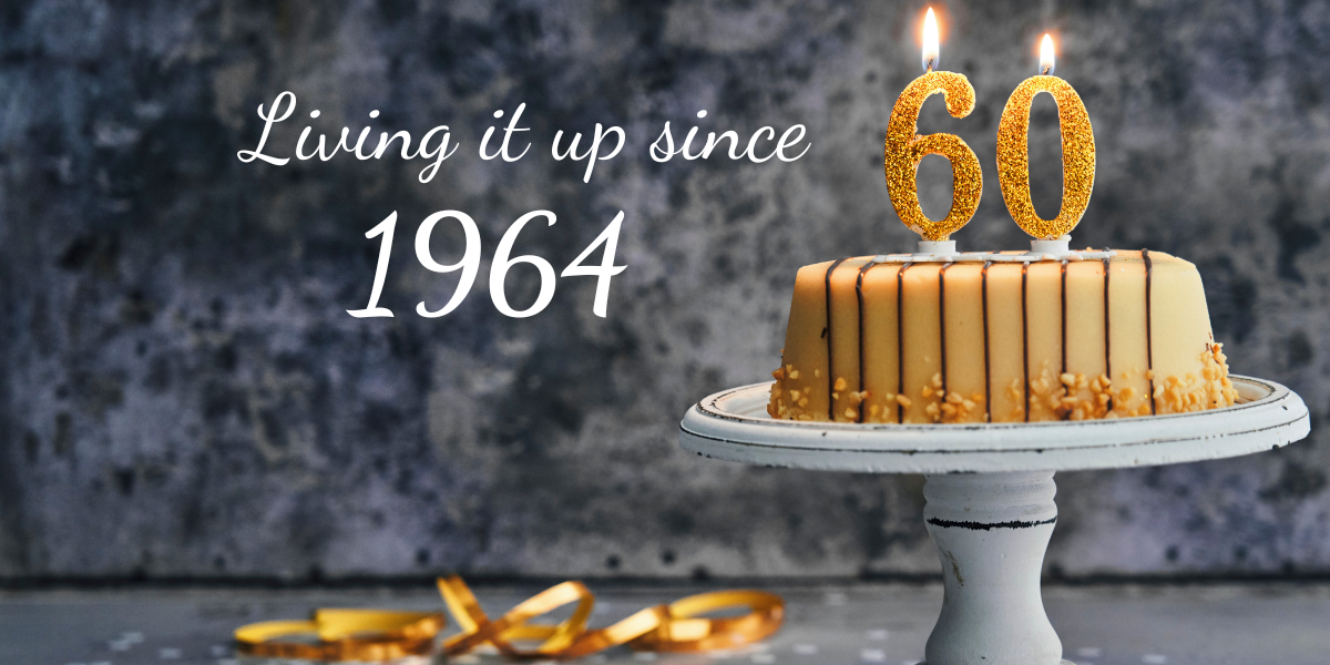 Mottled gray background. On the far right, a pale yellow frosted birthday cake on a cake plate with two lit gold candles shaped like "60." On the bottom of the photo, some curled gold ribbon. To the left is copy reading, "Living it up since 1964."